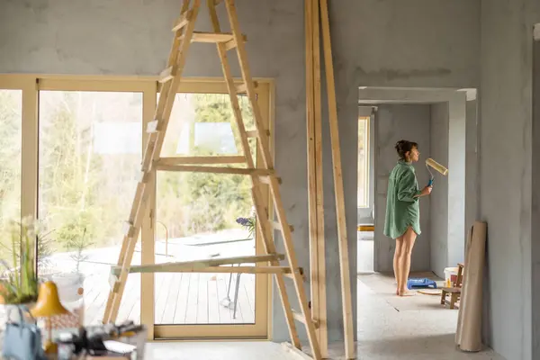 若い女性は新しい家を修理しながら壁を塗ります ペイントローラーで立って外を見る ホームリフォームと修理コンセプトのクリエイティブプロセス ロイヤリティフリーのストック画像