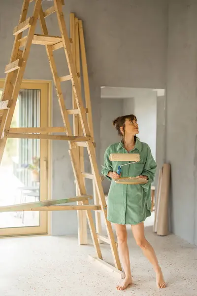 若い女性は新しい家を修理しながら壁を塗ります ペイントローラーで立って外を見る ホームリフォームと修理コンセプトのクリエイティブプロセス ストック写真