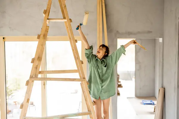 家の修理の過程でペイントローラーと一緒に梯子の上に幸せに立っている若いかわいい女性の肖像画 ホームリフォームと修理コンセプトのクリエイティブプロセス ストック写真