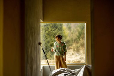 Genç bir kadın yeni bir evi tamir ederken duvarları boyuyor. Boya rulosuyla dikilip dışarı bakıyor. Yaratıcı ev yenileme ve onarım süreci