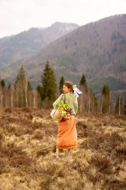 Pembe elbiseli genç bir kadın, bir çanta dolusu renkli çiçekle dağlardaki güzel bir çayırda yürüyor. Güzellik kavramı ve doğada tasasız yaşam tarzı.