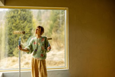 Genç bir kadın yeni bir evi tamir ederken duvarları boyuyor. Boya rulosuyla dikilip dışarı bakıyor. Yaratıcı ev yenileme ve onarım süreci