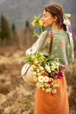 Pembe elbiseli genç bir kadının portresi. Elinde bir çanta dolusu renkli çiçeklerle dağlardaki güzel bir çayırda duruyor. Güzellik kavramı ve doğada tasasız yaşam tarzı.