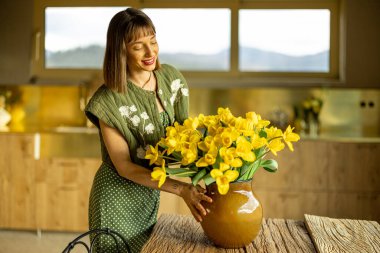 Zarif kadın, evdeki ahşap masada, şık evini dekore etmek için, bir vazo dolusu sarı çiçeği vazoya yerleştiriyor.