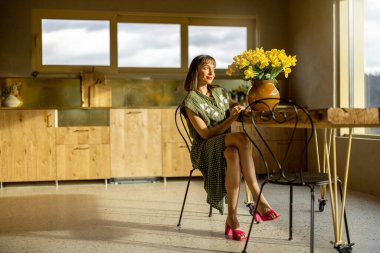 Genç bir kadın, şık ve ferah bir evde çiçeklerle süslenmiş yemek masasında rahatça oturur. Dağlarda bir evin iç kısımlarında.