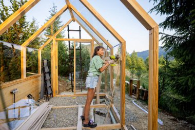 Dişi marangoz, arka bahçesinde ahşap bir serayı monte etmek için matkap kullanıyor. Günlük kıyafetler ve iş eldivenleri giyiyor. Doğa ile çevrili bir DIY projesiyle meşgul.