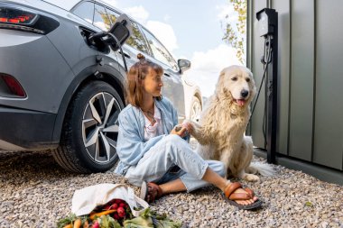 Küçük beyaz köpeğiyle birlikte oturan genç bir kadın, çantasında taze yiyeceklerle elektrikli araçla eve geldi. Mutlu ve sürdürülebilir yaşam tarzı, refah, evcil hayvanlarla arkadaşlık.