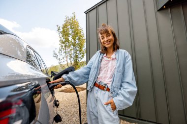Evinin yakınındaki elektrikli araca şarj aleti takan genç bir kadın. Modern teknolojiler, EV arabalar ve sürdürülebilir yaşam tarzı kavramı