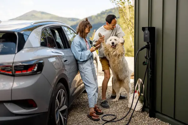 Evlerinin yanında sevimli beyaz köpekleri ve modern elektrikli arabaları olan genç bir çiftin portresi. Aile ve refah kavramı