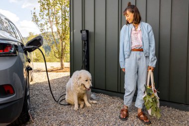 Genç bir kadın, sevimli köpeği ve alışveriş çantasıyla eve giderken elektrikli arabasıyla modern evine geldi. Sürdürülebilir yaşam tarzı, refah ve evcil hayvanlarla arkadaşlık kavramı