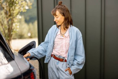 Evinin yakınındaki elektrikli araca şarj aleti takan genç bir kadın. Modern teknolojiler, EV arabalar ve sürdürülebilir yaşam tarzı kavramı