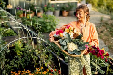 Genç bir kadın elinde taze sebzeler ve renkli çiçeklerle dolu bir sepet tutarken gülümsüyor ve bakımlı bahçesinde bir anın tadını çıkarıyor.