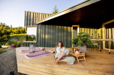 A woman relaxes on a wooden deck with a peaceful mountain view, surrounded by nature. The calm atmosphere of this mountain retreat offers an escape from the hustle and bustle of daily life. clipart