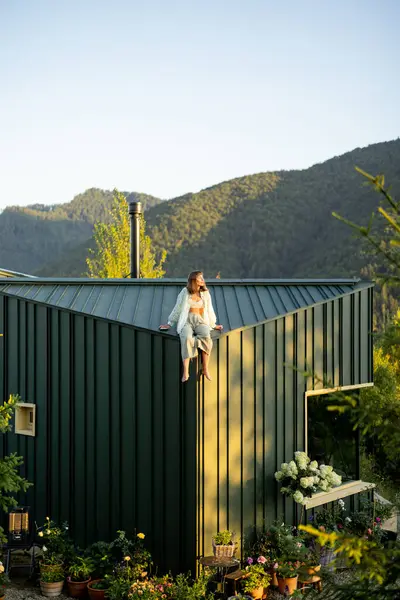 stock image A woman enjoys a tranquil morning on a mountain cabin rooftop, surrounded by lush nature and stunning views, embodying rest, relaxation, and a deep connection with the wilderness.