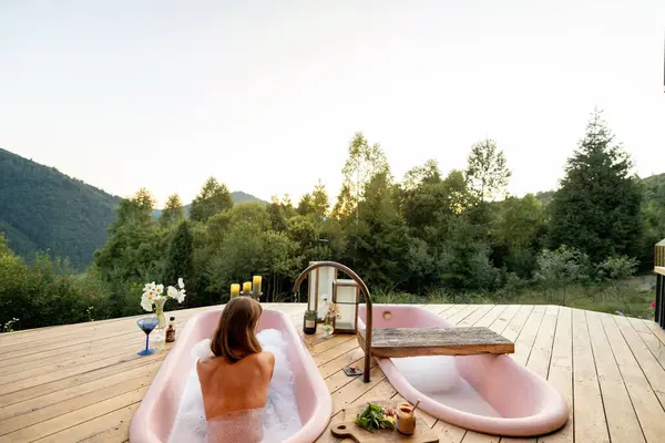 stock image A serene moment of relaxation as a woman enjoys a bubble bath in a pink bathtub on a wooden deck, surrounded by the tranquil beauty of nature at sunset