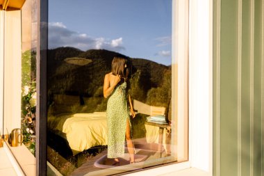 Bir kadın içeride duruyor, saçını düzeltiyor, yansıması modern bir evin geniş penceresine mükemmel bir şekilde yansıyor. Çevresindeki doğa yansıtılır, iç kısmı dışarıdaki manzarayla harmanlanır.