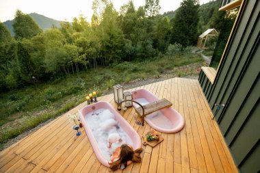 Dağlardaki yemyeşil bir ormana tepeden bakan ahşap bir güvertede duran pembe küvette rahatlatıcı bir köpük banyosu. Rahatlamak ve doğaya dalmak için mükemmel bir kaçış.