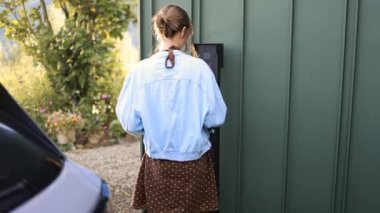 Genç bir kadın bir şarj istasyonunda elektrikli arabasına şarj prizi bağlıyor. Sahne, sürdürülebilir yaşamı ve elektrikli araçlara yönelik modern değişimi yansıtıyor.