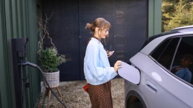 Gözlük takan ve şık giyinen genç bir kadın, elektrikli arabasından çıkıp bir şarj istasyonuna bağlıyor. Görüntü, eko-bilinçli yaşamın günlük rutinini yakalar