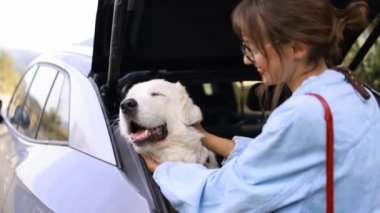 Bir kadın, elektrikli bir arabanın bagajında oturan büyük beyaz köpeğini kucaklıyor ve bir yolculuğa hazırlanıyor. Bu yürek ısıtan an, evcil hayvan ve sahibinin arasındaki bağı ve sevgiyi yakalar.
