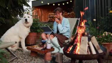 Bir kadın, tabletinde bir film izleyerek sıcak bir akşam geçirir, yanında sadık köpeği ile birlikte, sakin bahçedeki çıtırdayan ateşin sıcaklığında otururken.
