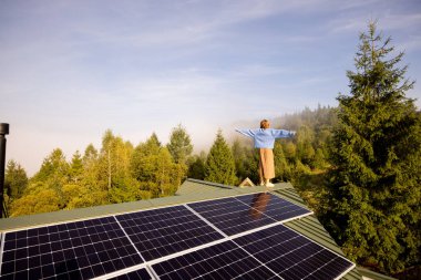 A woman stands on a solar-powered rooftop, arms outstretched, embracing the freedom and sustainability of off-grid living amidst a serene forested landscape clipart