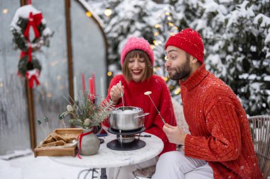 Erkek ve kadın fondüyle romantik bir akşam yemeği yerken, aynı zamanda güzel dekore edilmiş karlı arka bahçede masanın başında oturuyorlar. Genç bir aile kış tatilini dışarıda kutluyor.