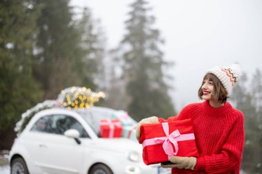 Kırmızı kazaklı ve şapkalı mutlu kadın portresi, hediyelerle süslenmiş bir araba ve dağlarda Noel ağacı ile seyahat ederken hediye kutusuyla ayakta duruyor. Mutlu kış tatili kavramı