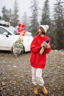 Hediyelerle süslenmiş arabayla seyahat ederken telefonu kullanan bir kadın ve dağlarda Noel ağacı. Doğada yeni yılı kutlayan bir kız.