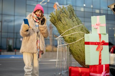 Genç, neşeli bir kadın, alışveriş arabasıyla alışveriş merkezinin otoparkında arabanın yanında dururken telefonla konuşuyor.