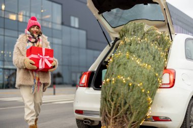 Kışın giyinmiş neşeli genç bir kadın alışveriş merkezinin yanında hediye kutusuyla duruyor ve önünde Noel ağacı var. Kış tatili için alışveriş kavramı