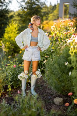 Bir kadın canlı bir çiçek bahçesinin ortasında, açık mavi lastik botlarının içine tıkıştırılmış taze beyaz papatyalarla güvenle duruyor.