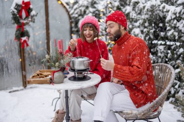 Erkek ve kadın fondüyle romantik bir akşam yemeği yerken, aynı zamanda güzel dekore edilmiş karlı arka bahçede masanın başında oturuyorlar. Genç bir aile kış tatilini dışarıda kutluyor.