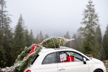 Çatıda Noel ağacı olan beyaz bir araba, arka kapıda çelenk ve karlı havada dağ yolunda sunulan hediyeler. Yeni yıl anlayışı ve tatil hazırlığı.