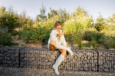 A woman enjoys a peaceful moment in her garden, scrolling through her phone as the morning sun bathes the scene in warm light. Her outdoor space reflects a balance of modern living and nature clipart