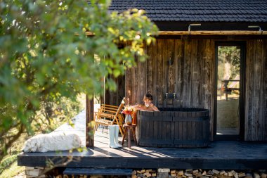 Bir kadın açık havada, ahşap bir banyoda, sıcak bir içecek yudumlarken rahatlıyor. Çiçekler ve doğal ışıkla kırsal alan huzurlu ve huzurlu bir atmosferi yakalar.
