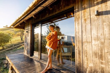 Turuncu bir kazakla ahşap bir güvertede duran kadın, altın sabah güneşinin tadını çıkarıyor. Camdaki yansımalar sakin ve sakin bir atmosferi yakalıyor.