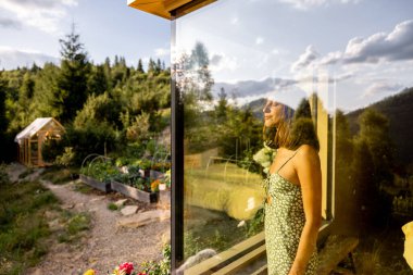 Modern bir evde, geniş bir pencerenin yanında duran bir kadın, yansıması açık hava yeşiliyle kusursuz bir şekilde harmanlanmış.