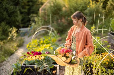 Gün batımında çiçek açan bir bahçede elinde bir sepet taze koparılmış çiçek tutan genç bir kadın. Doğayla mükemmel bir uyum anı.