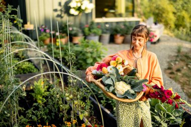 Genç bir kadın elinde taze sebzeler ve renkli çiçeklerle dolu bir sepet tutarken gülümsüyor ve bakımlı bahçesinde bir anın tadını çıkarıyor.