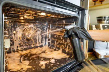 A close-up of a hand in a black glove spraying cleaning solution inside a dirty oven. The foam cleaner is applied to remove grease and grime, ensuring a spotless and hygienic appliance clipart