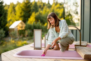 A woman prepares an outdoor bathtub by placing fresh flowers in the water, setting the stage for a relaxing spa experience amidst the serene mountain landscape