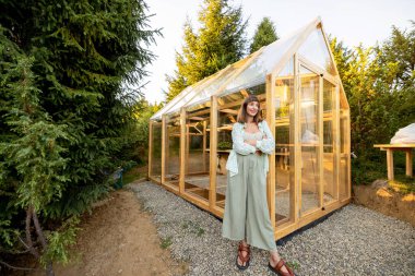 Smiling woman with a basket of fresh vegetables stands outside a greenhouse, radiating happiness and pride in her homegrown produce clipart