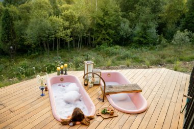 Dağlardaki yemyeşil bir ormana tepeden bakan ahşap bir güvertede duran pembe küvette rahatlatıcı bir köpük banyosu. Rahatlamak ve doğaya dalmak için mükemmel bir kaçış.