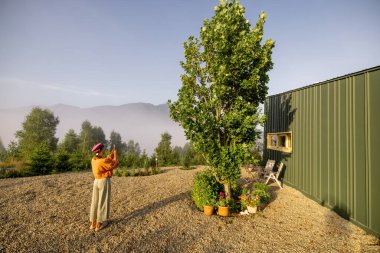 A woman takes a photo of the serene morning mist enveloping the mountains, standing beside a modern home surrounded by nature, highlighting the tranquility of countryside living clipart