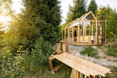 A wooden greenhouse under construction in a lush backyard surrounded by tall trees. The scene captures the early stages of a DIY project in an eco-friendly environment, highlighting sustainable living clipart