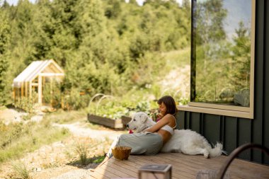 Bir kadının ve sadık köpeğinin verandada dinlenip doğal manzaranın güzelliğini içlerine çektikleri sakin bir sahne. Camdaki yansıma kompozisyona derinlik katıyor.