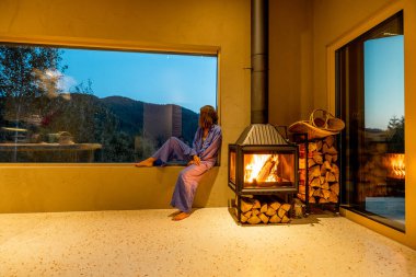 Woman sitting on a windowsill near a fireplace, enjoying the twilight mountain view. Warm interior with rustic decor blends nature with comfort clipart