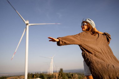 Geniş kolları olan, rüzgar enerjisini kucaklayan, büyük rüzgar türbinlerinin önünde duran yenilenebilir enerji ve sürdürülebilirliği sembolize eden kişi.