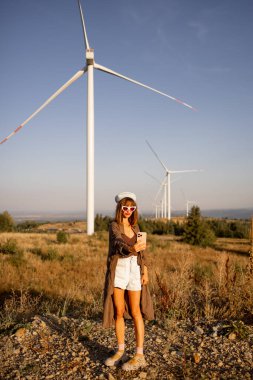 Tarz sahibi bir kadın, modern moda ve sürdürülebilir enerjinin kesişiminde manzaralı bir kırsal alanda devasa rüzgar türbinlerinin önünde selfie çekiyor.